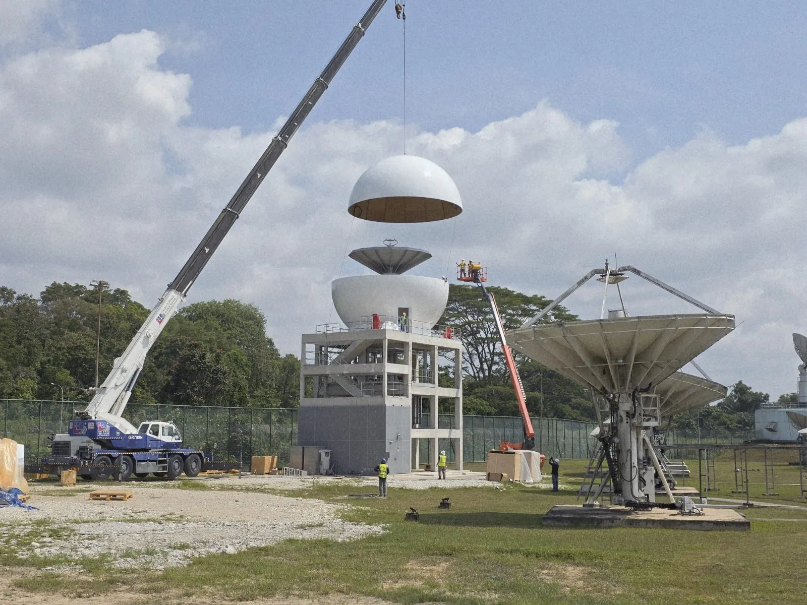 A crane lowers the top of a radome onto a LEO antenna on its tower while crews work on site